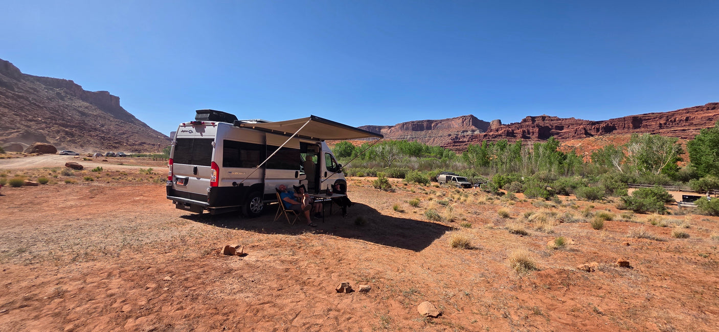 A white RV rental with an extended awning offers shade to two people relaxing in a vast red rock desert.