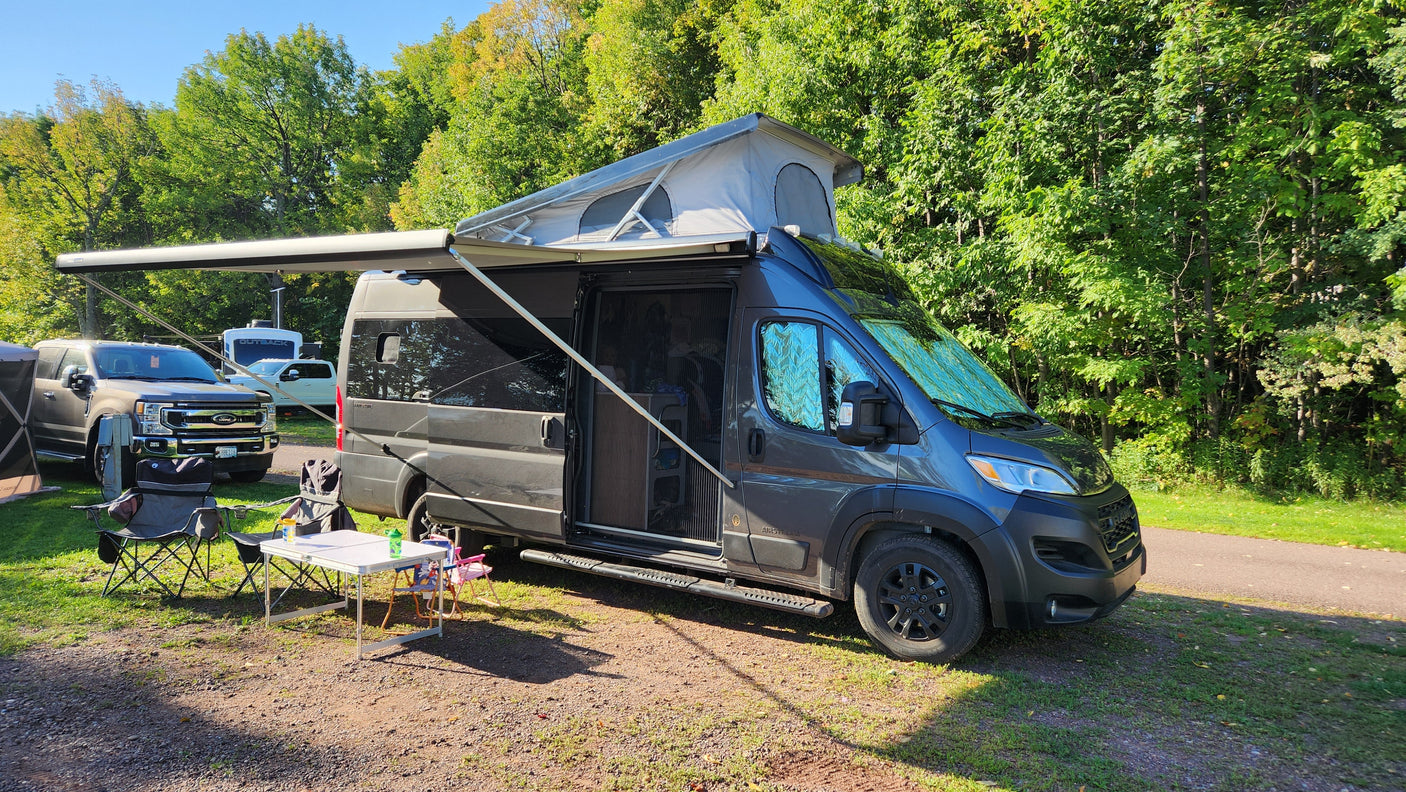 An RV rental, a dark grey camper van with its pop-top and awning out, is set up at a sunny campsite with chairs and a table.