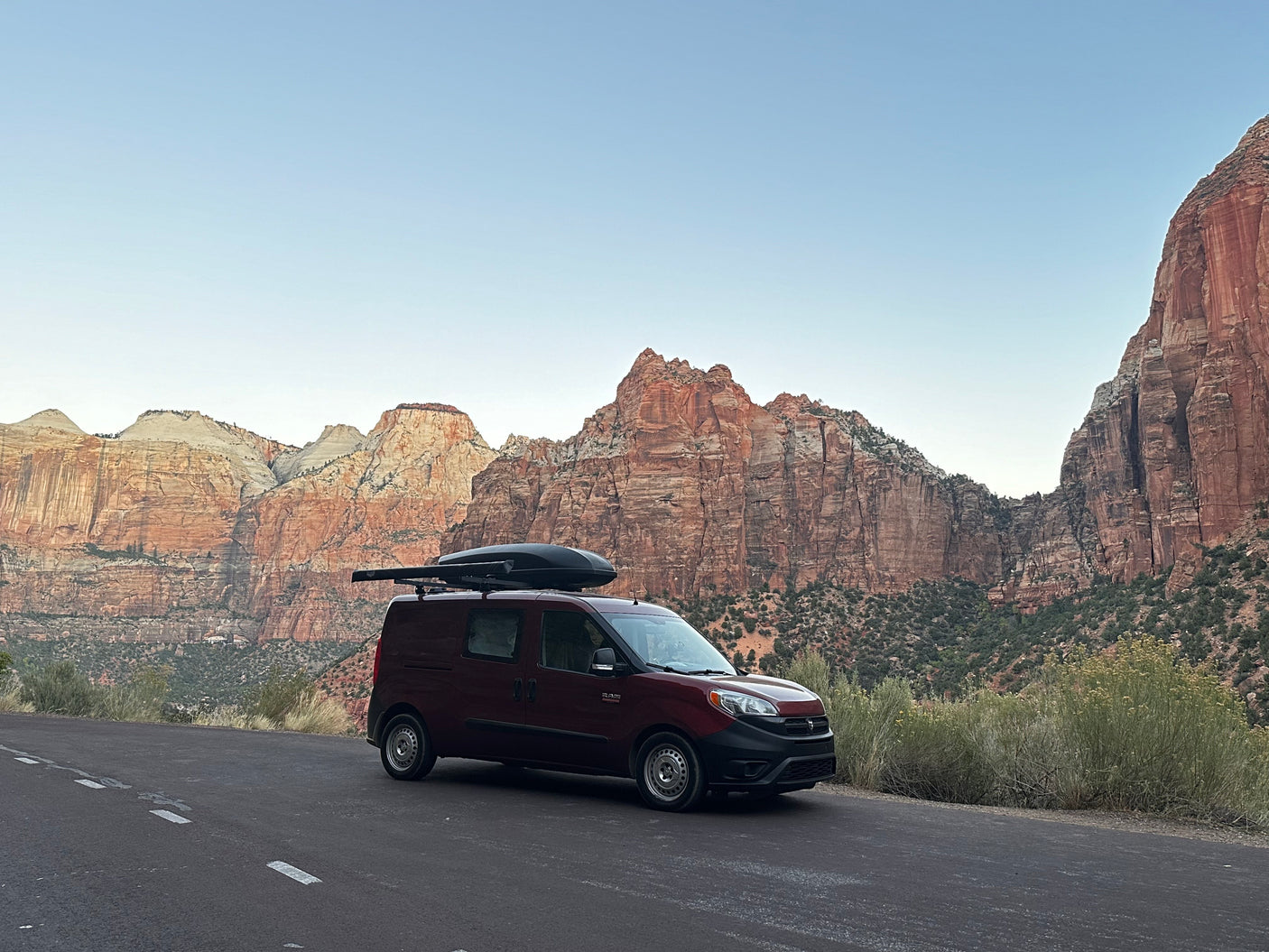 A dark red RV rental van with a roof cargo box is parked on a road, facing majestic red rock mountains.
