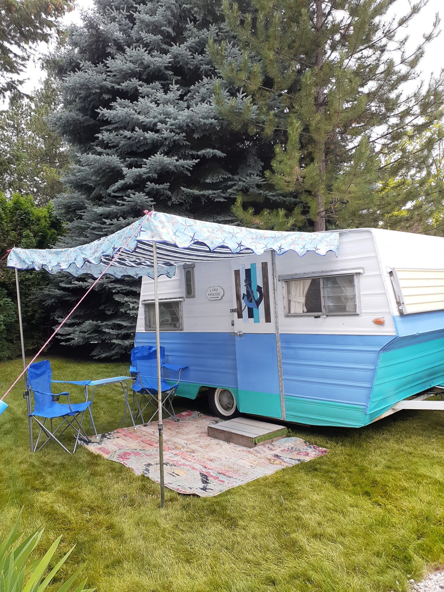 Relaxing RV rental scene: a retro blue & teal camper with an awning, chairs, and rug on green grass.