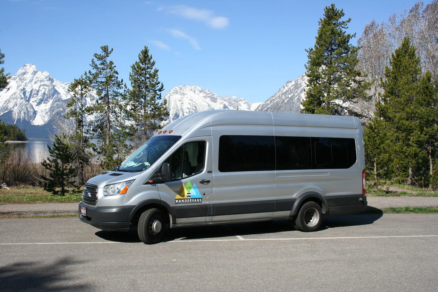 Silver RV rental van by a scenic lake, with snow-capped mountains and pine trees in the background.