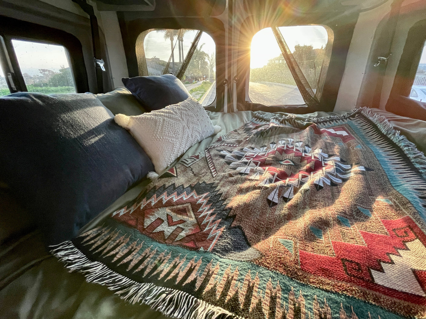 Cozy RV rental bed with pillows & patterned blanket, bright sun shining through windows, offering a relaxing road view outside.