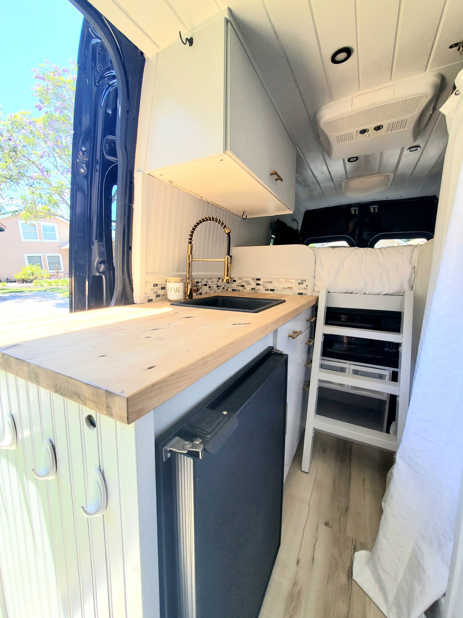 Compact RV rental interior featuring a stylish kitchen with gold faucet, fridge, and built-in ladder to the bed, ready for travel.