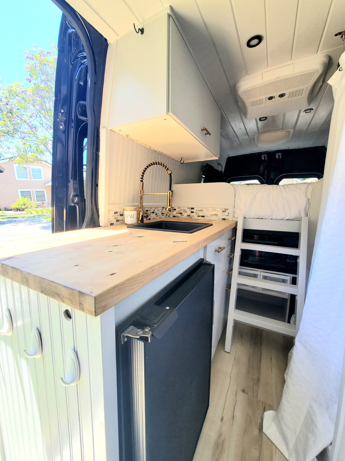 Compact RV rental interior featuring a stylish kitchen with gold faucet, fridge, and built-in ladder to the bed, ready for travel.
