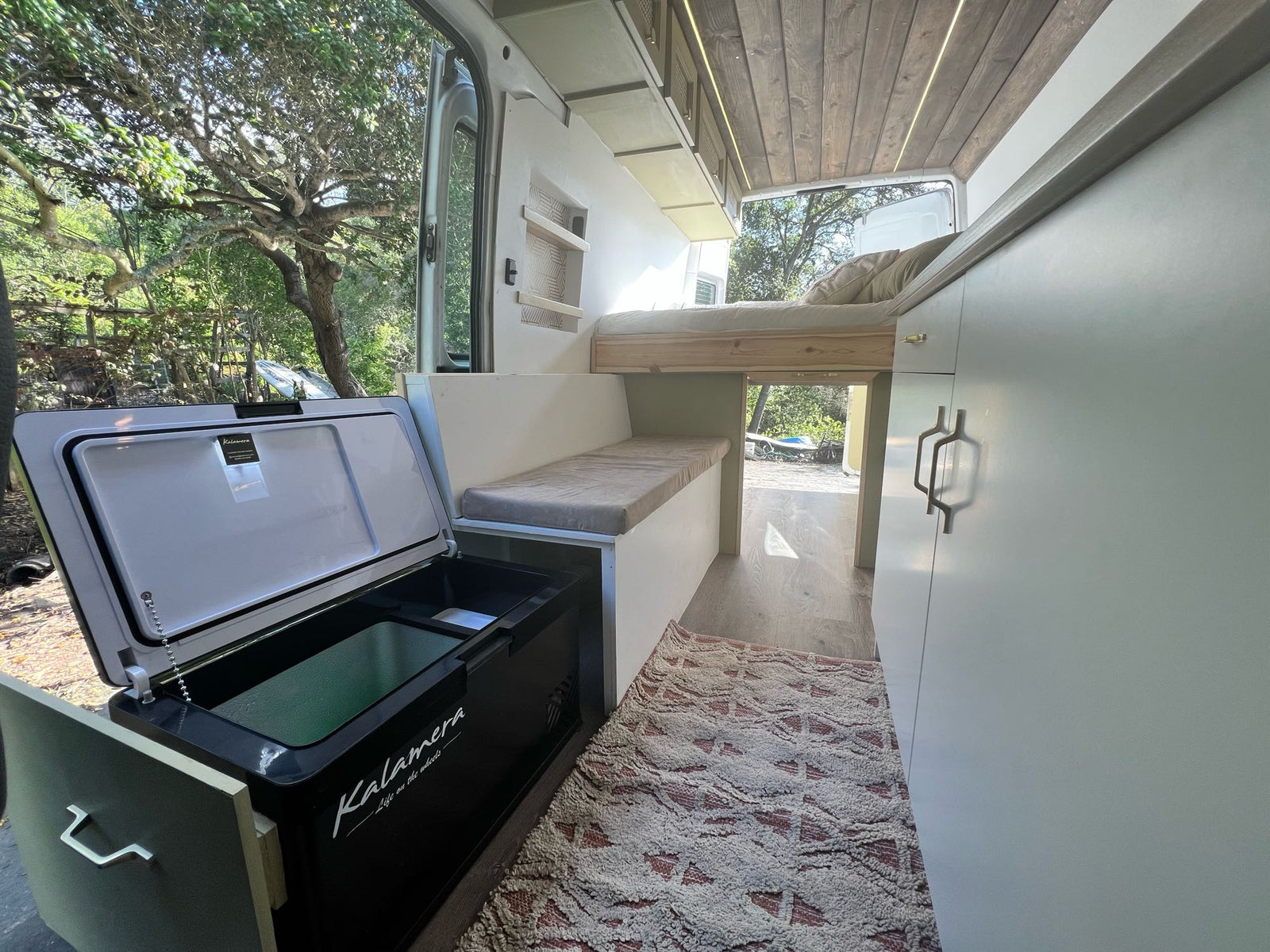 Inside a modern RV rental: a cozy raised bed, bench seating, and an open fridge with outdoor nature views.