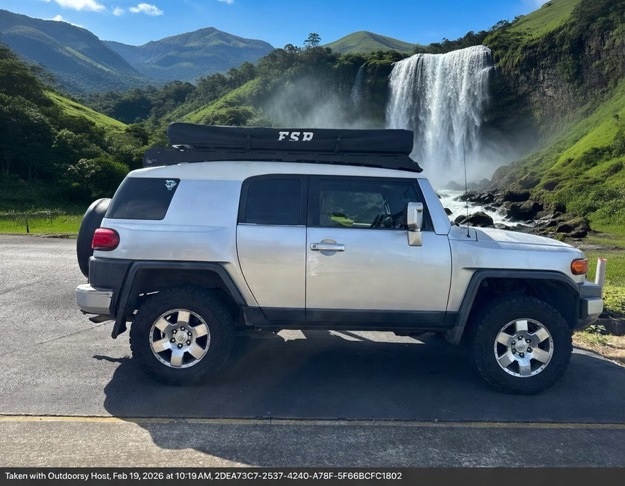RV rental adventure! Silver Toyota FJ Cruiser with a rooftop tent by a majestic waterfall and green mountains.