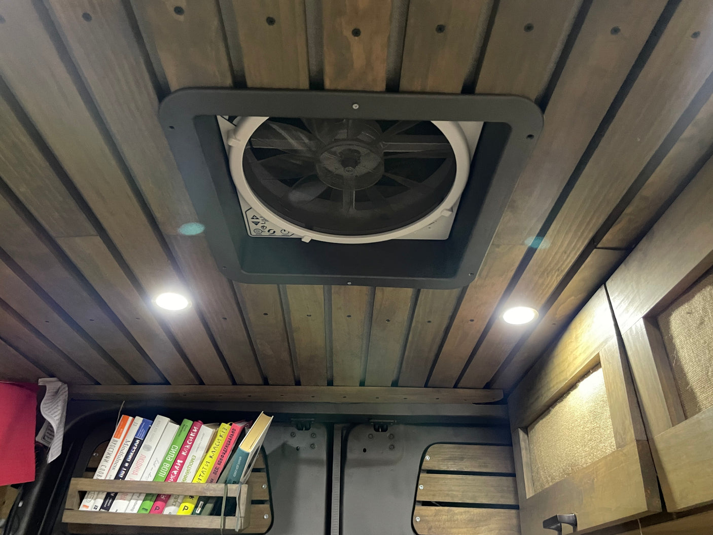 RV rental interior: Wooden ceiling with fan, lights, and a bookshelf filled with books. A cozy van life setup.