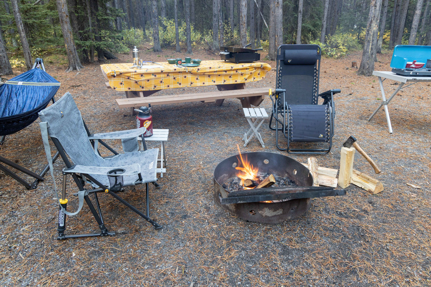 Inviting RV rental campsite featuring a fire pit, hammock, cooking area on a picnic table, and comfy chairs amidst pine trees.