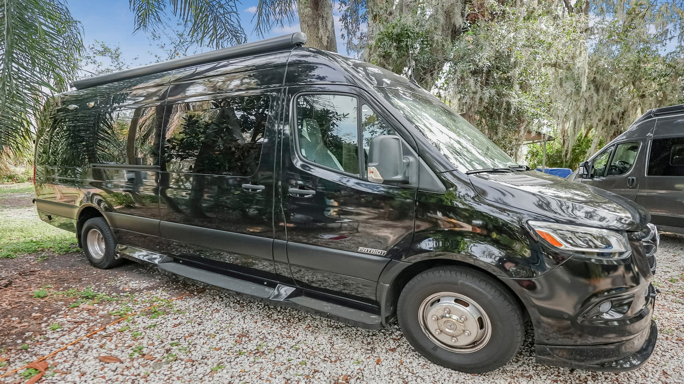 This black Patriot Sprinter RV rental is parked on gravel, featuring a rooftop awning and sleek design amidst lush trees.