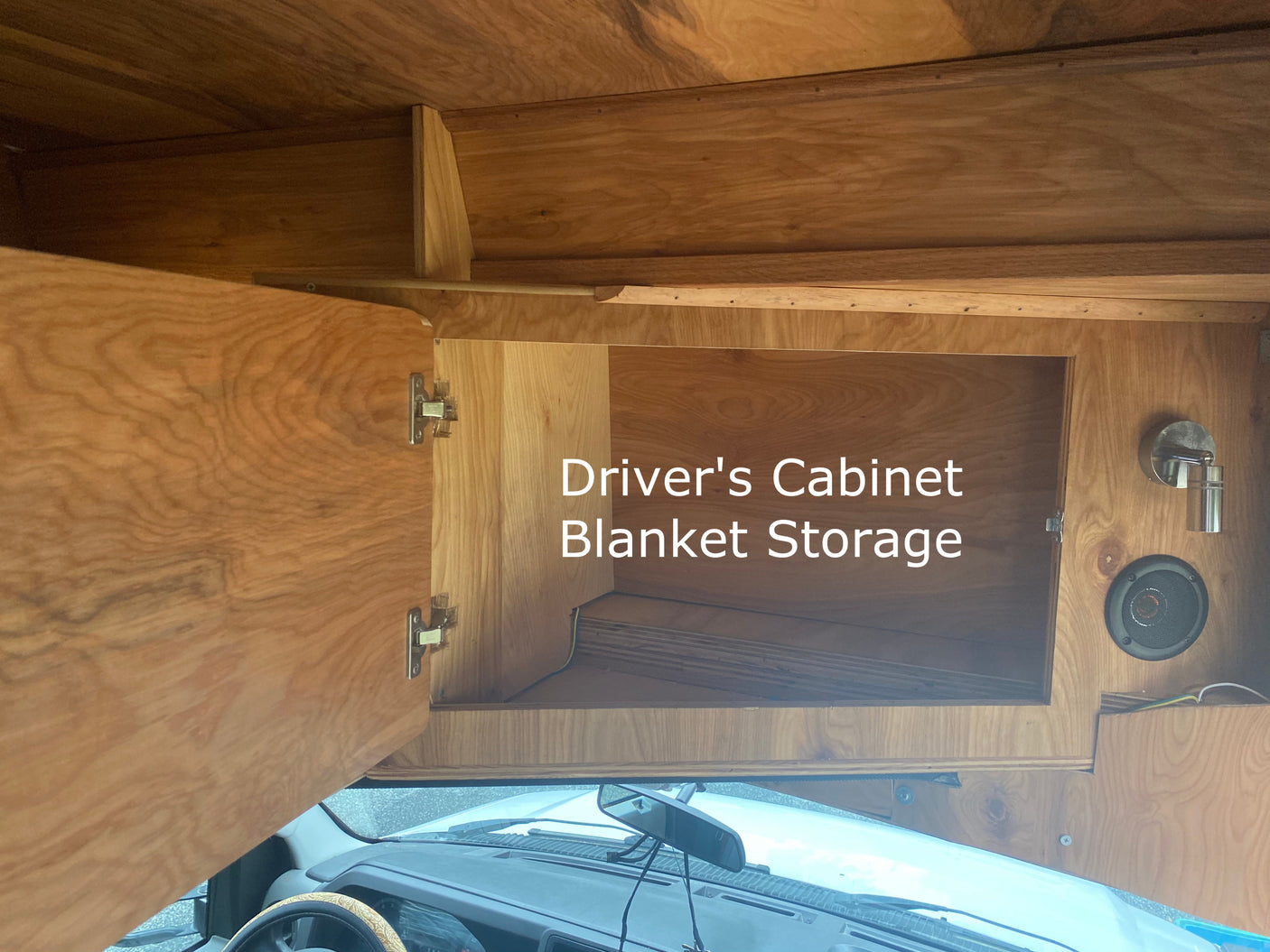 Inside an RV rental: view of a custom-built wooden driver's cabinet for blanket storage, open to show the vehicle's dashboard.