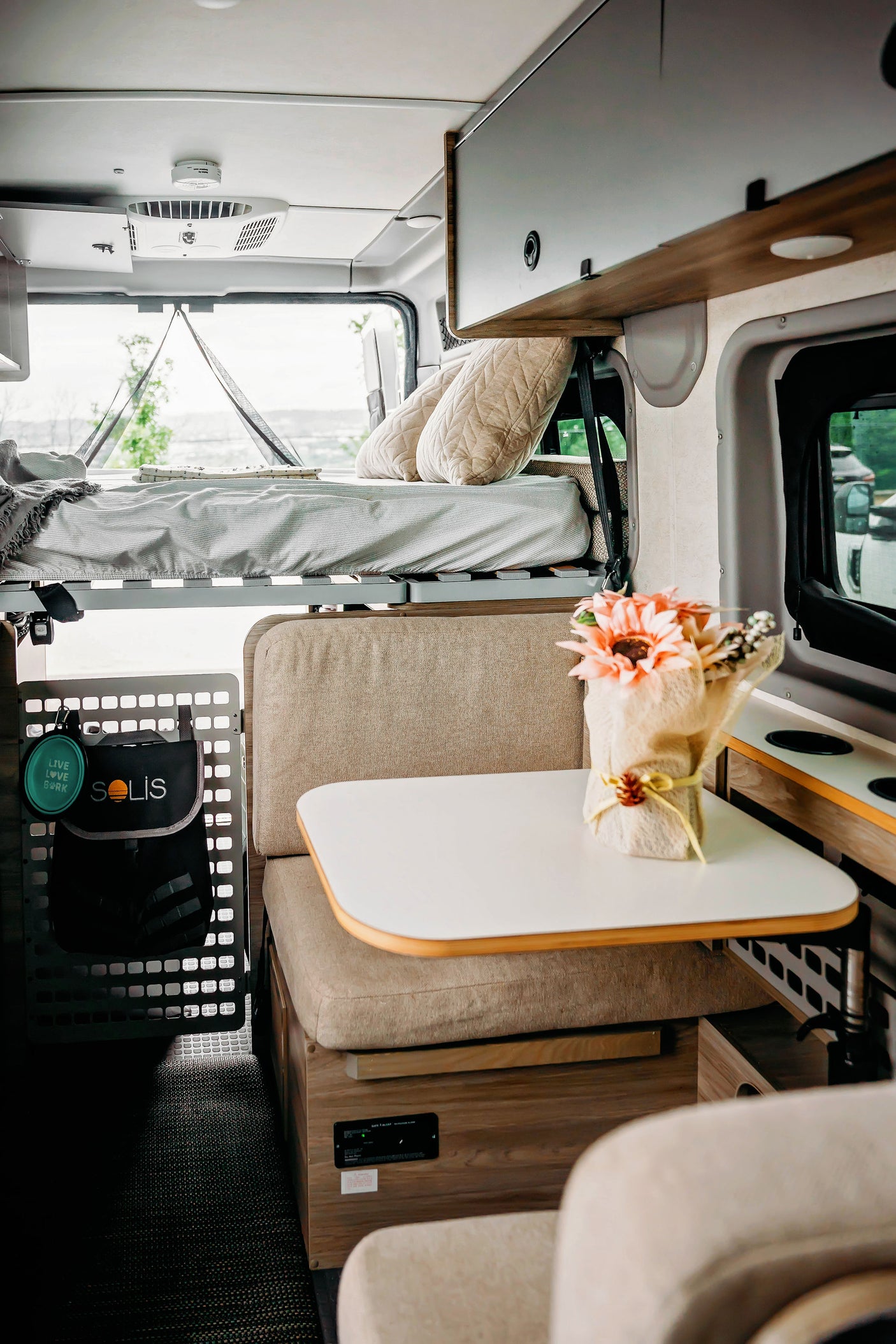 Modern rv rental interior: elevated bed, comfy dinette with table, overhead storage, and dog travel accessories visible.