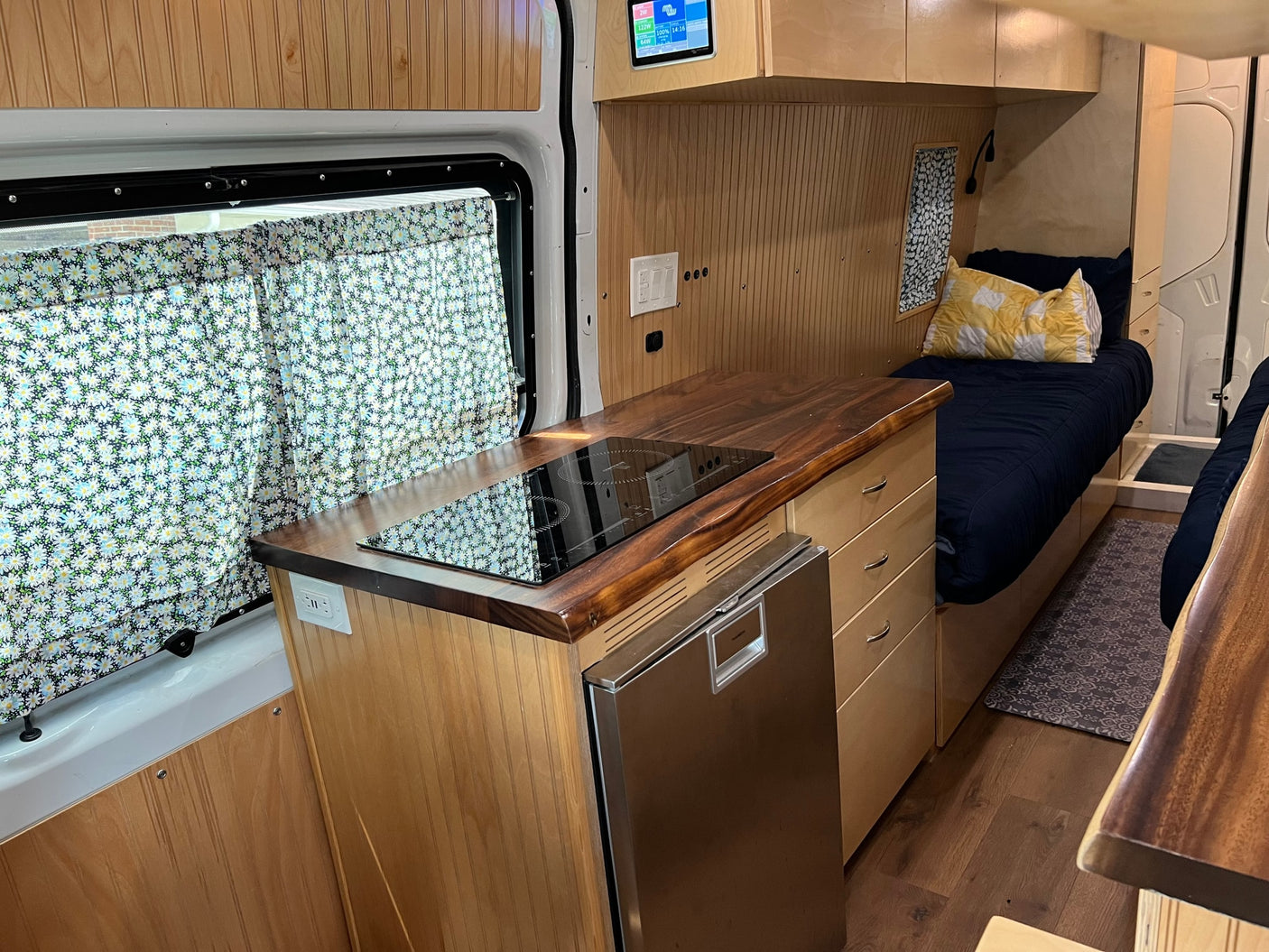 Compact RV rental interior featuring a kitchen with induction cooktop, fridge, and cozy bed area with daisy curtains.