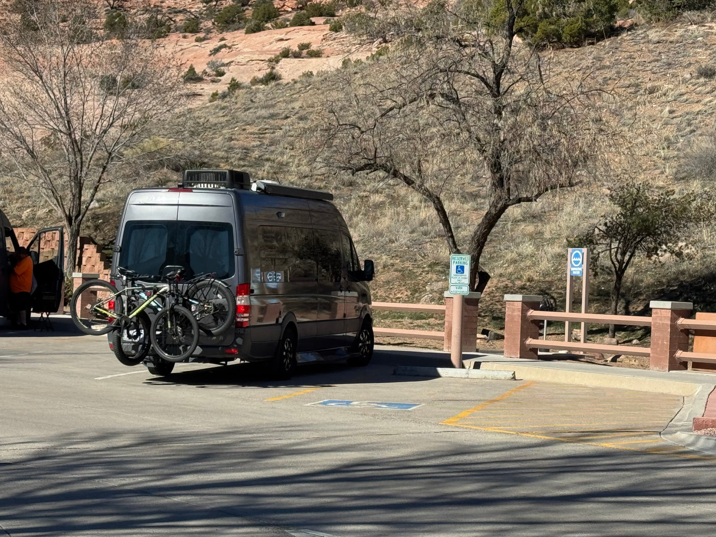 relaxing outside a gray rv rental camper van at a sunny campsite with drinks on a table.|Dark gray RV rental with bikes on a snowy desert road. Dramatic winter clouds loom over snow-dusted hills.|A gray rv rental van with bikes on a rack