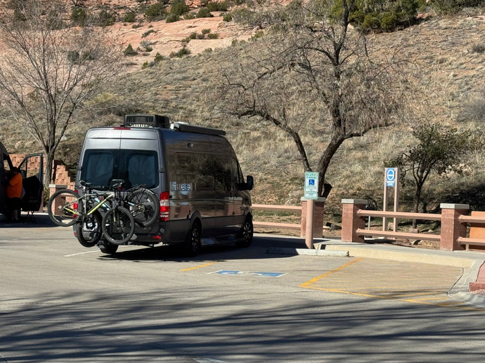 relaxing outside a gray rv rental camper van at a sunny campsite with drinks on a table.|Dark gray RV rental with bikes on a snowy desert road. Dramatic winter clouds loom over snow-dusted hills.|A gray rv rental van with bikes on a rack