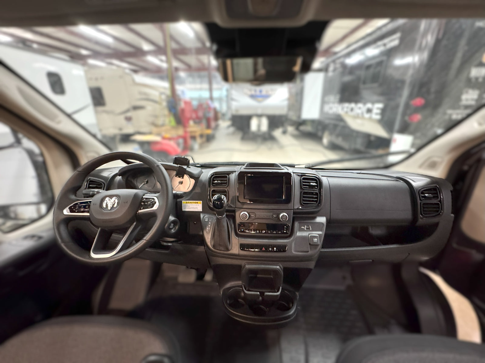 View from inside a Ram van: dashboard, steering wheel. Blurry background shows RVs and trucks in a warehouse. Ideal for rv rental.
