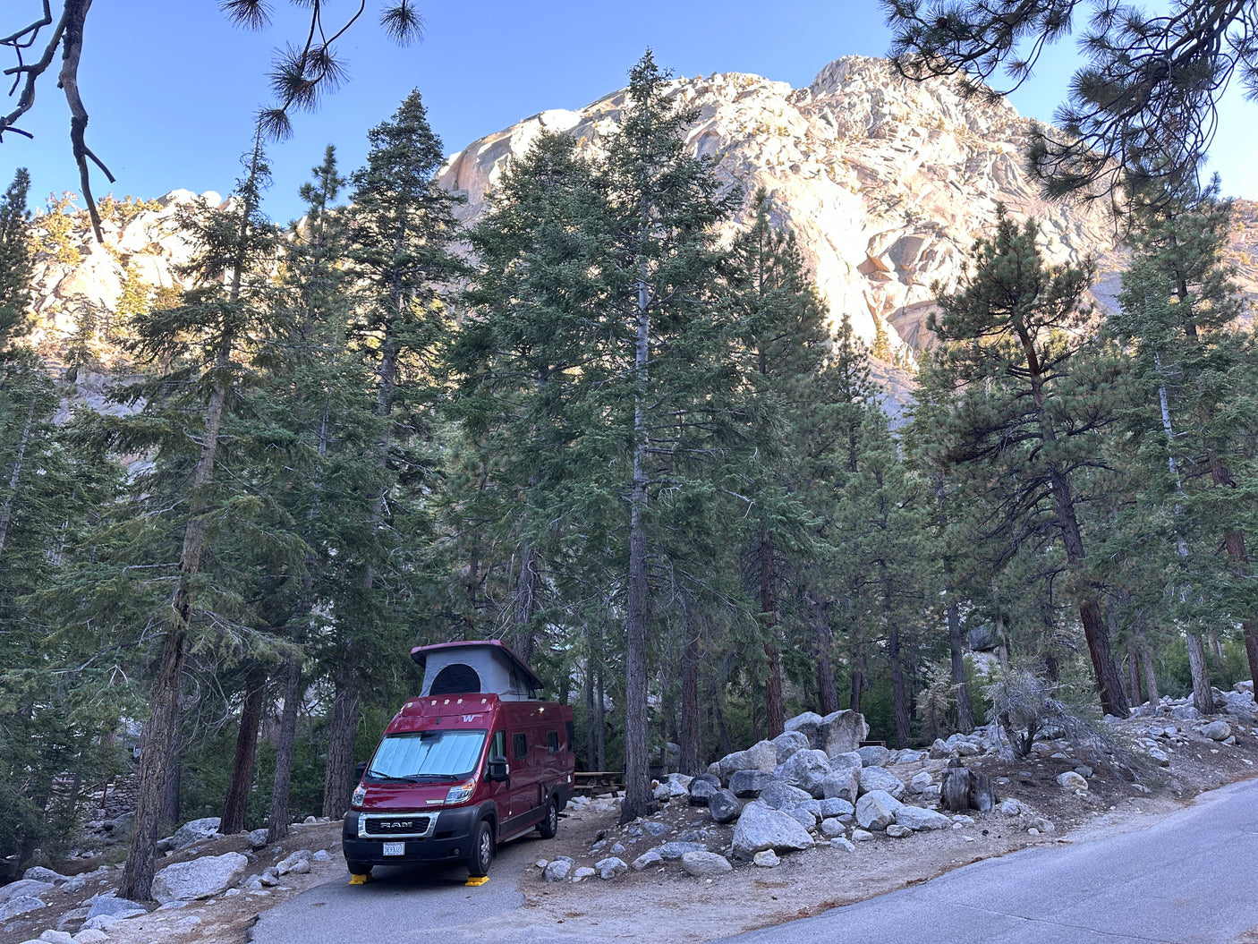 Red RV rental camper van at a wooded campsite, framed by trees against a sunny granite mountain.