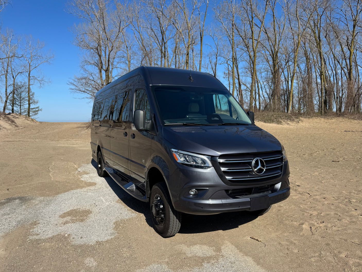 Explore with this dark gray Mercedes Sprinter RV rental. Parked on a sandy path near water, with bare trees under a blue sky.