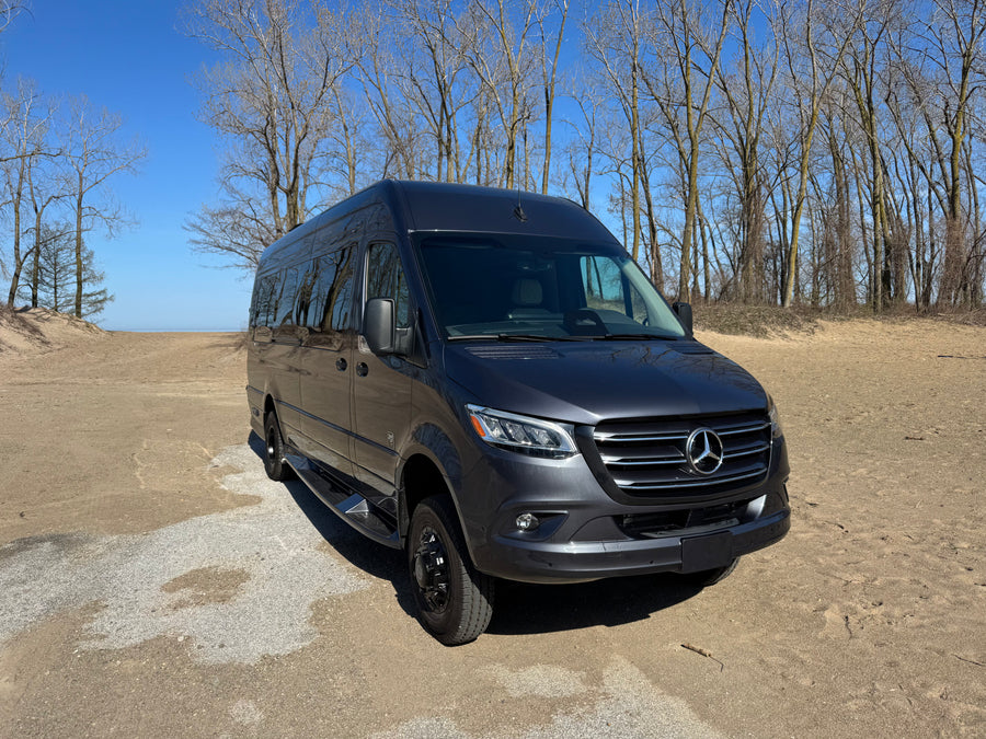 Explore with this dark gray Mercedes Sprinter RV rental. Parked on a sandy path near water, with bare trees under a blue sky.
