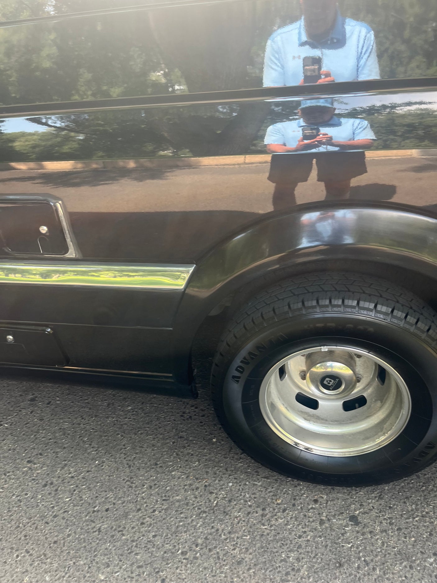 A shiny dark RV side, with its tire and reflections of a person & trees. Perfect for an RV rental.