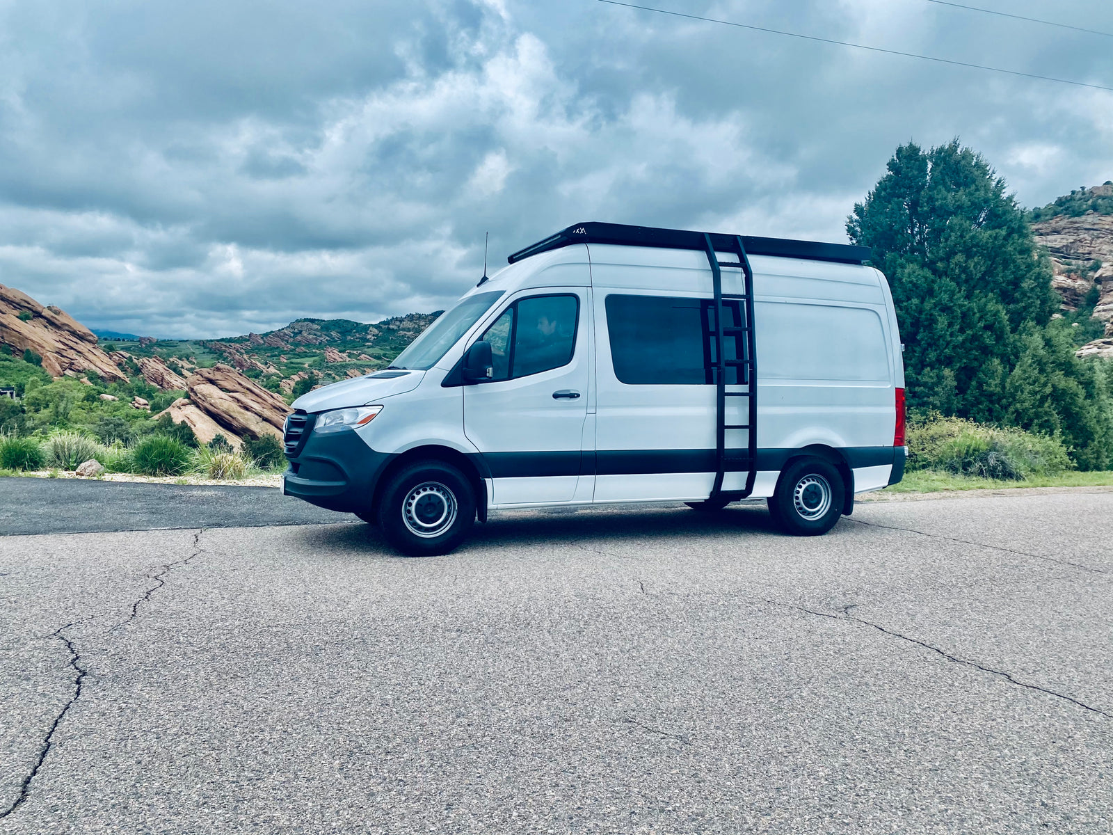Scenic RV rental, a white Sprinter van, parked on an asphalt road with red rocks and cloudy skies in the background.