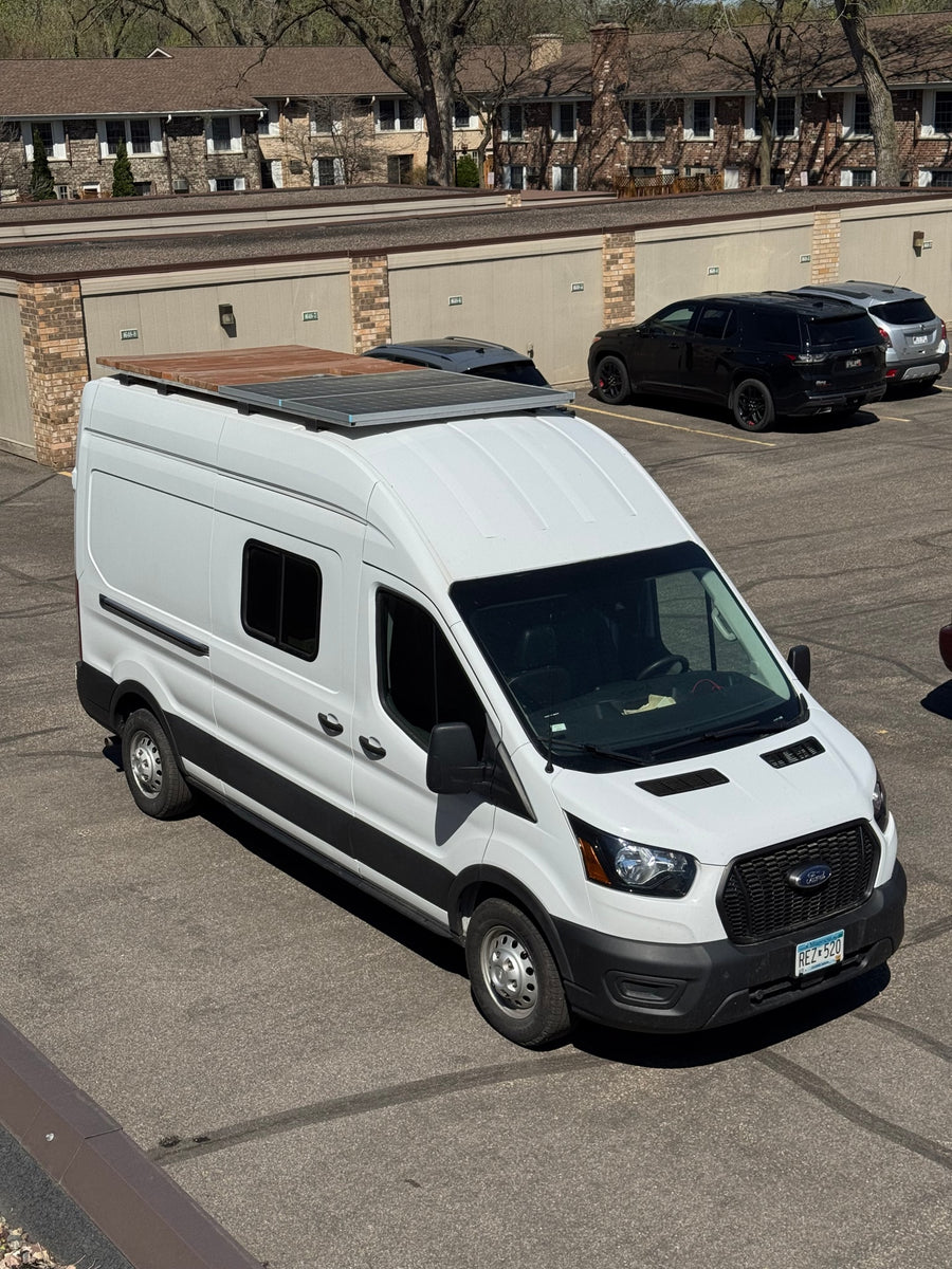 White Ford Transit camper van, possibly an rv rental, parked in a lot with solar panels and a roof deck.