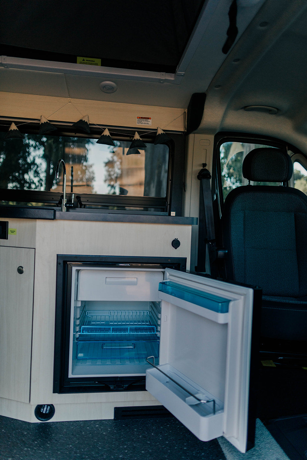 Inside this RV rental, a compact kitchen is ready: an open mini-fridge reveals its shelves, next to a sink with a window view of trees.