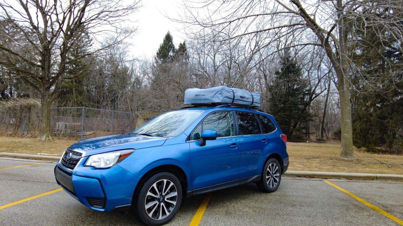 A blue Subaru SUV with a Thule rooftop tent, parked in a lot surrounded by bare trees. An option beyond standard RV rental.