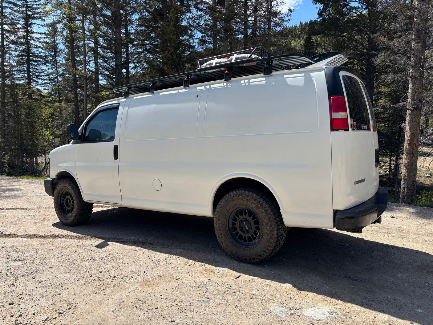 White camper van, equipped with a roof rack and off-road tires, parked on a forest dirt road. Ideal for an RV rental adventure.