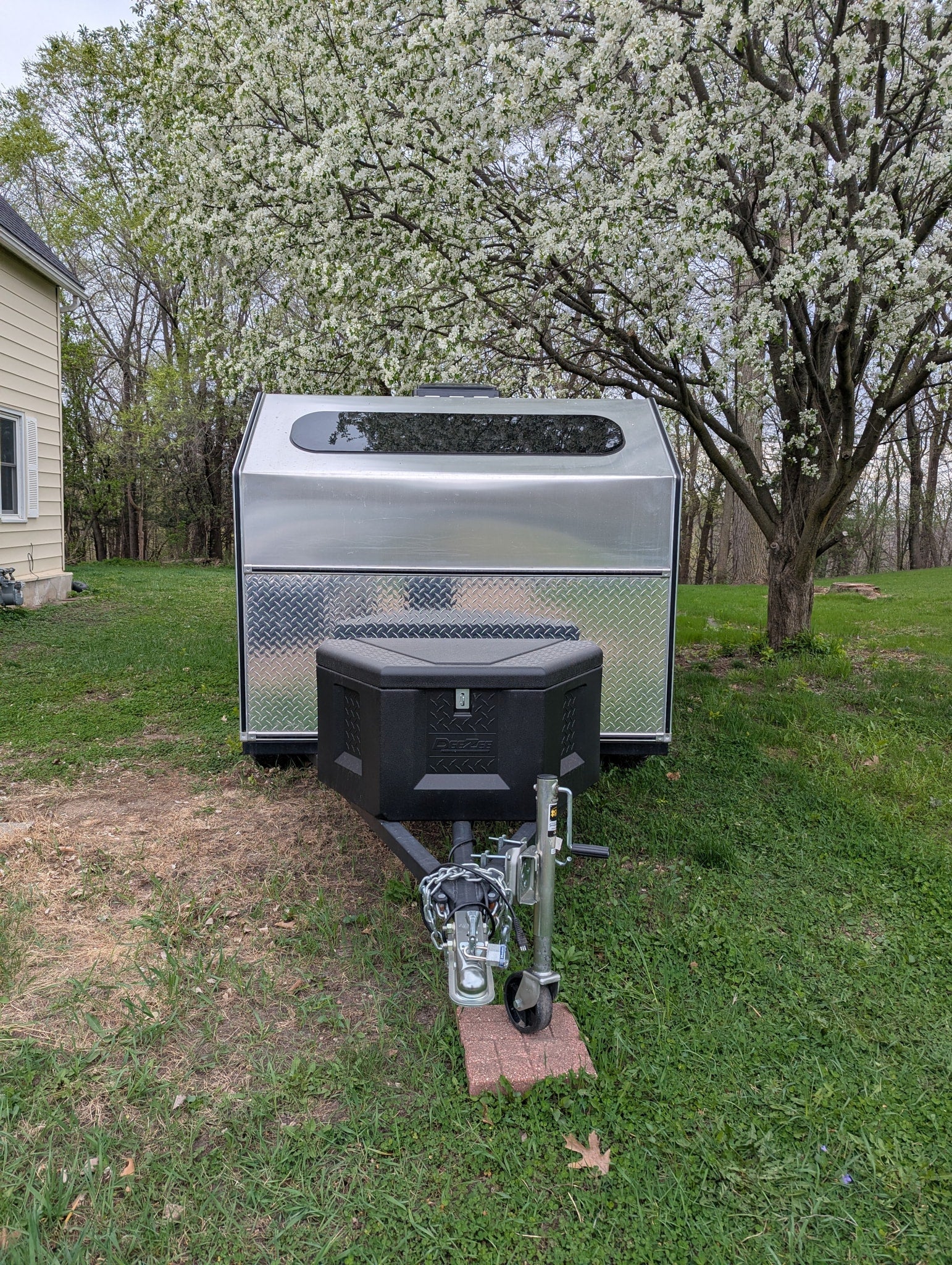 An aluminum teardrop camper, perfect for RV rental, is parked in a grassy yard under a white blooming tree.