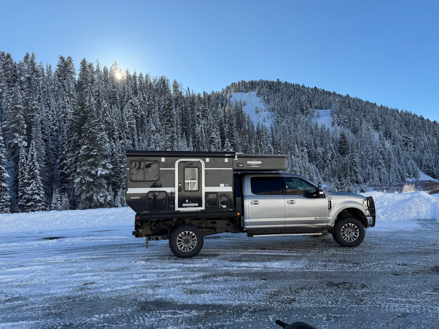 Silver truck with a black Four Wheel Campers pop-top camper, ready for an RV rental adventure, parked on an icy road in snowy mountains.