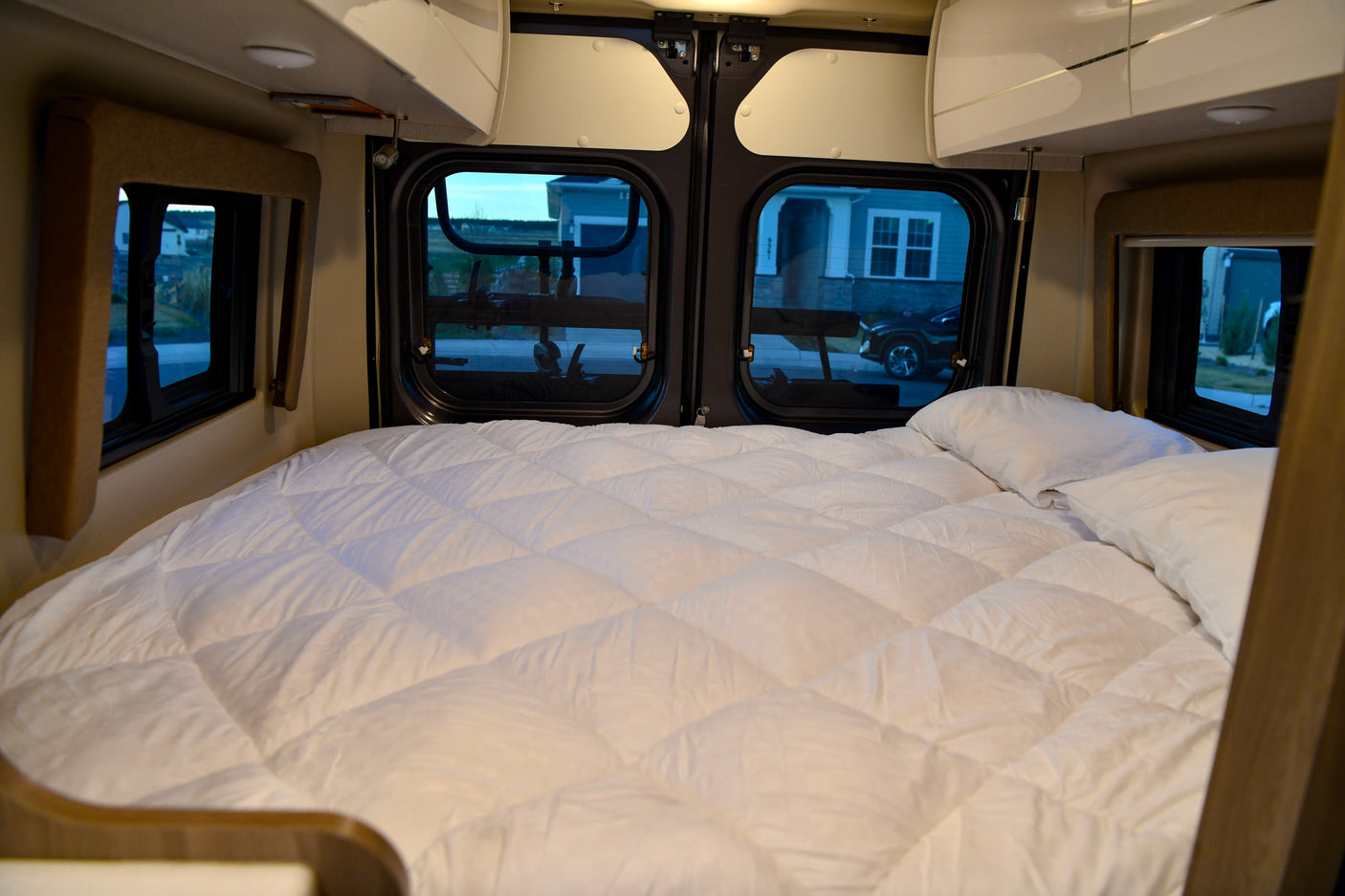 Plush white bed inside an rv rental camper van, offering comfy sleep with clear window views of the neighborhood.