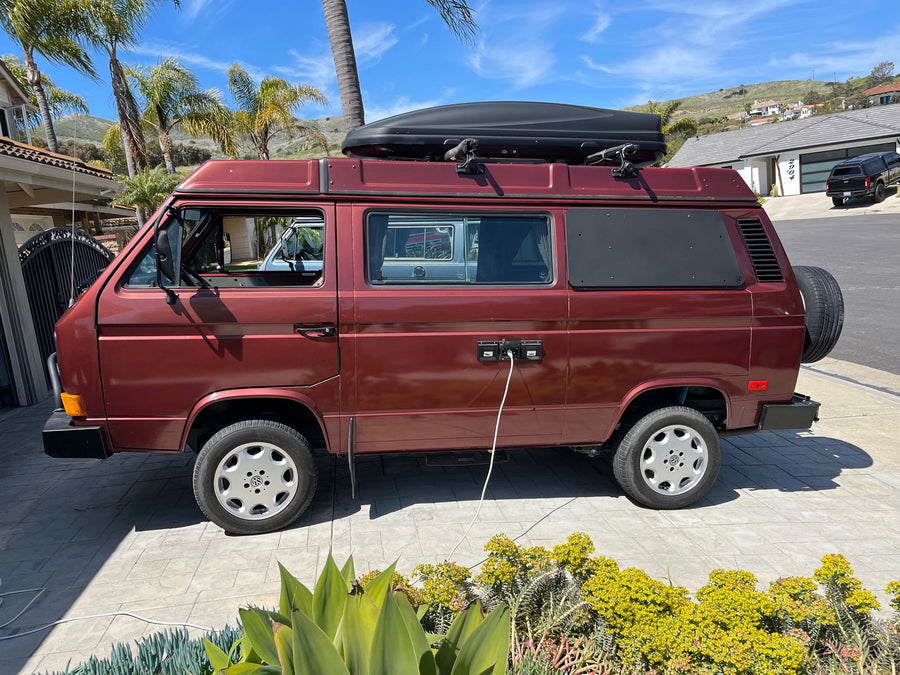 A classic red VW camper van, ideal for your next RV rental. It's parked on a sunny driveway, ready with a roof box and power.