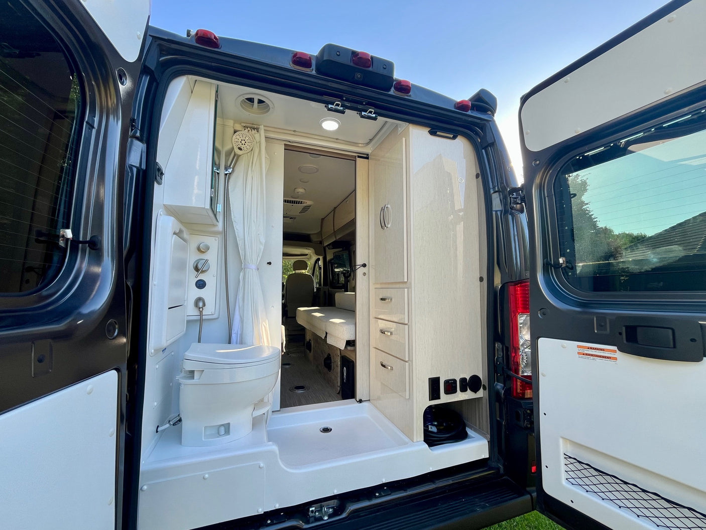 Rear view of open RV rental doors revealing a compact shower, toilet, and storage inside this modern camper van.