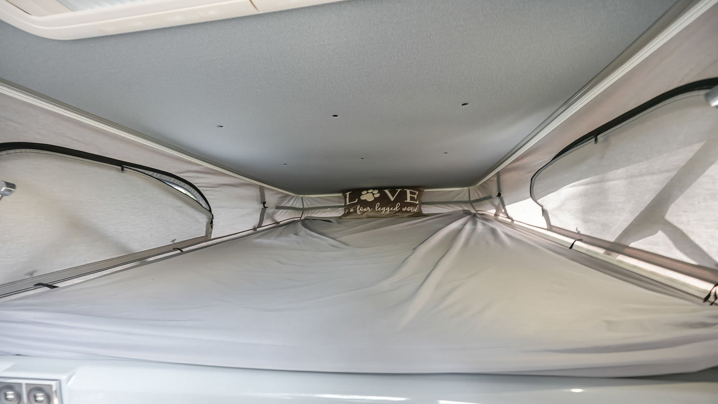 Inside a cozy pop-top RV rental, showcasing the raised ceiling, light gray interior, and a comfy sleeping area with a pet-themed pillow.