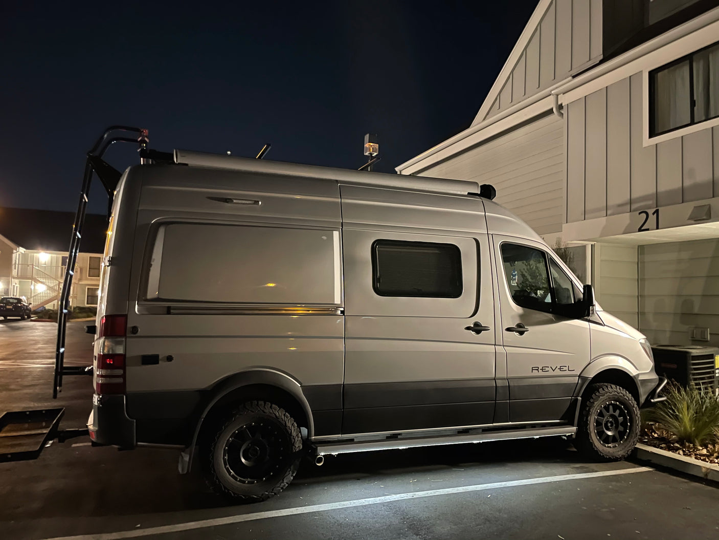 Night shot of a silver Winnebago Revel camper van with a rear ladder & rugged tires, great for RV rentals.