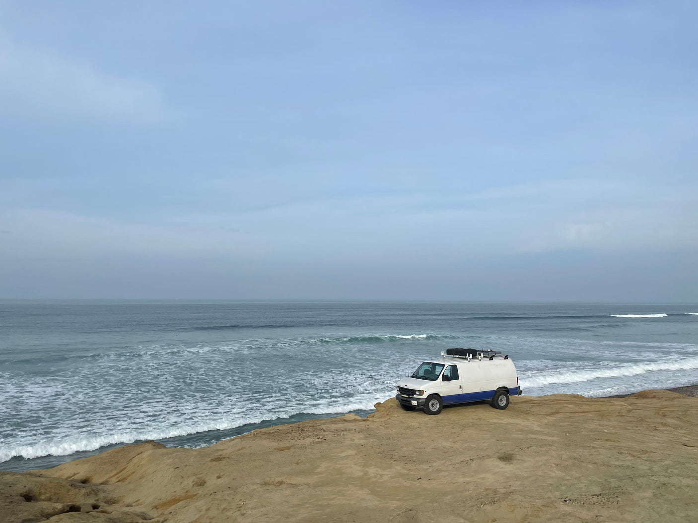 An RV rental, a white and blue van, is parked on a sandy cliff overlooking the ocean under cloudy skies.