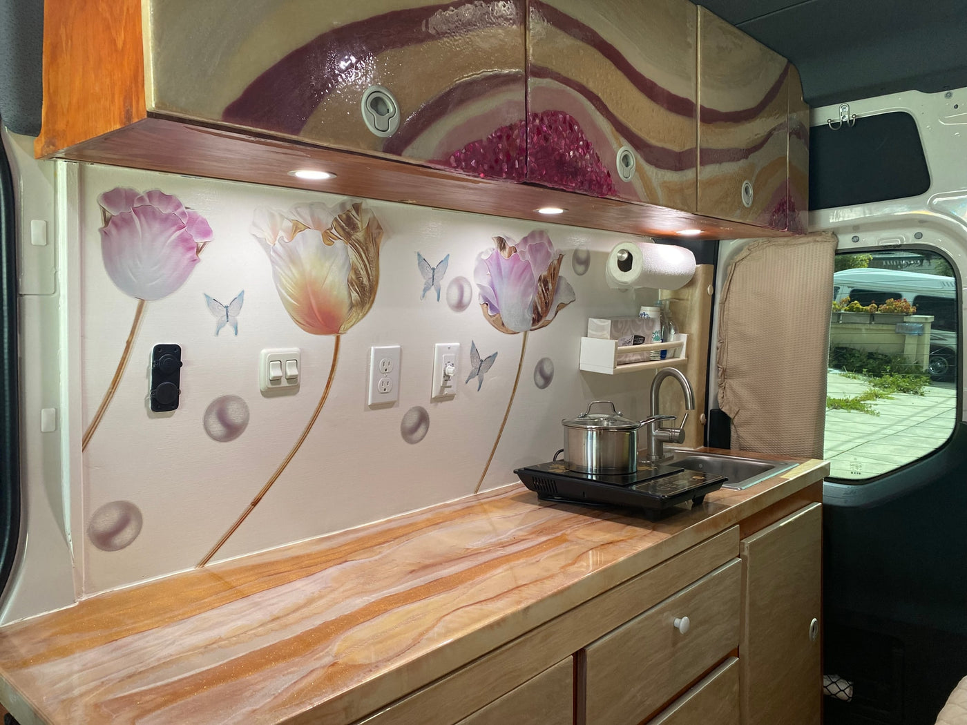 Experience this unique RV rental! Custom van interior featuring a vibrant kitchen with artistic decor, functional countertop, and cooking area. — photo 7