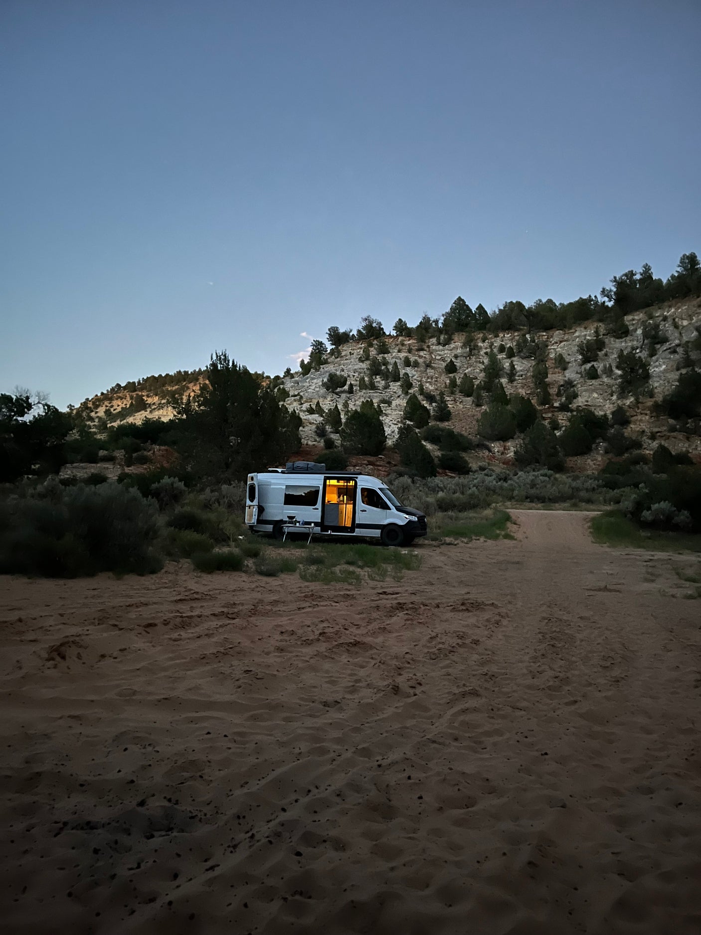 An RV rental camper van with interior lights on is parked at dusk on a sandy desert road near rocky hills, perfect for boondocking.