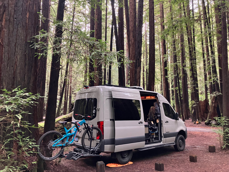 RV rental van parked in a serene redwood forest with bikes, ready for outdoor adventure and exploring nature.