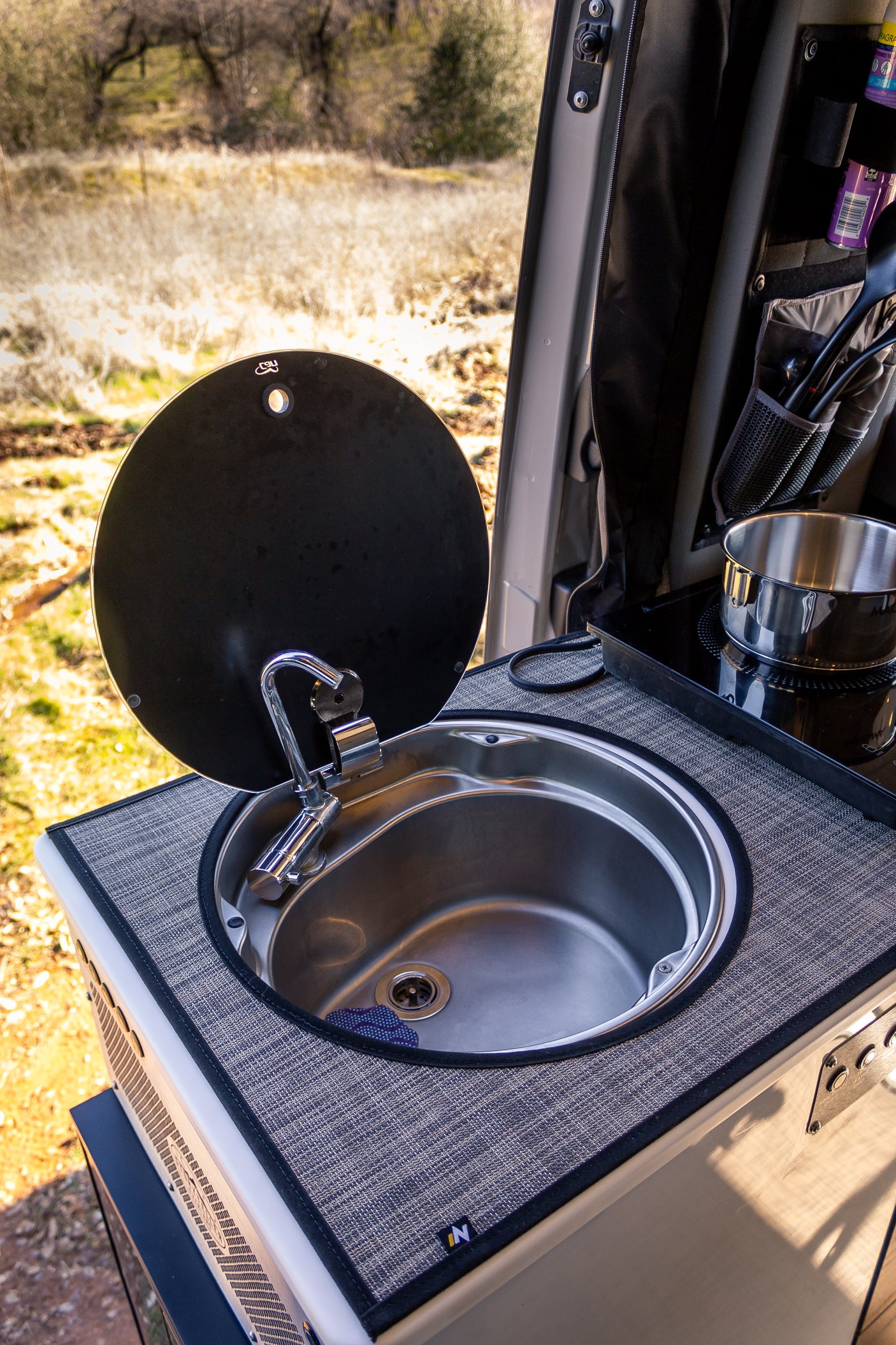 This RV rental's compact kitchen features a stainless steel sink with a faucet, a lift-up cover, and an induction cooktop with a pot, ready for meals in nature.