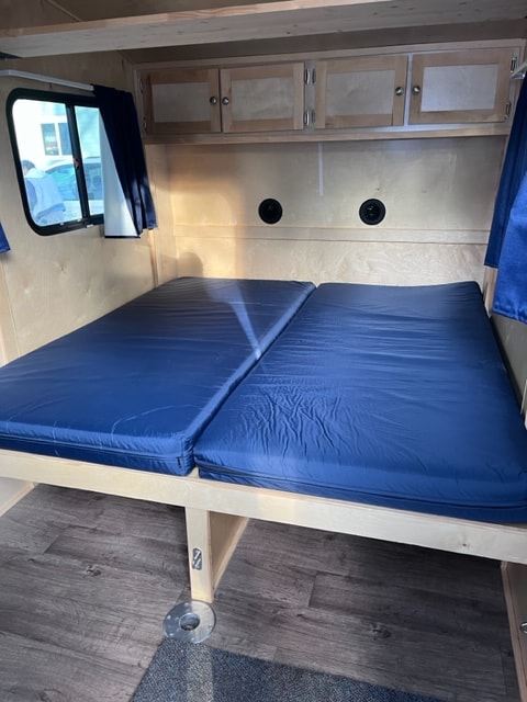 Inside a compact rv rental: two blue bunk beds on the right, light wood cabinets on the left, and a window below.