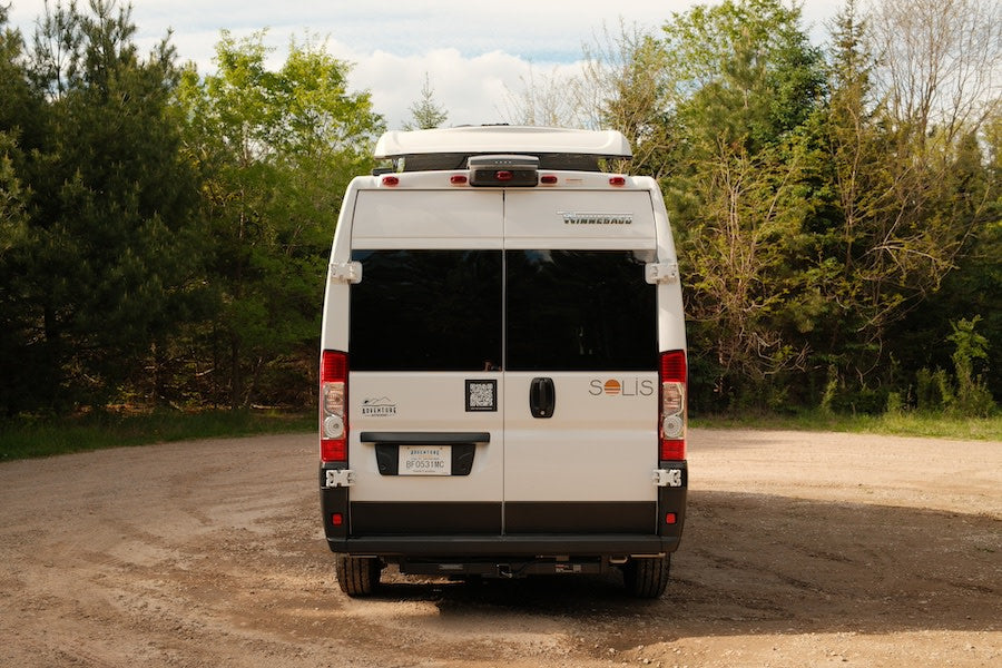 Rear of a white Winnebago Solis RV rental, parked on a dirt road in a wooded setting. Ready for your next adventure.