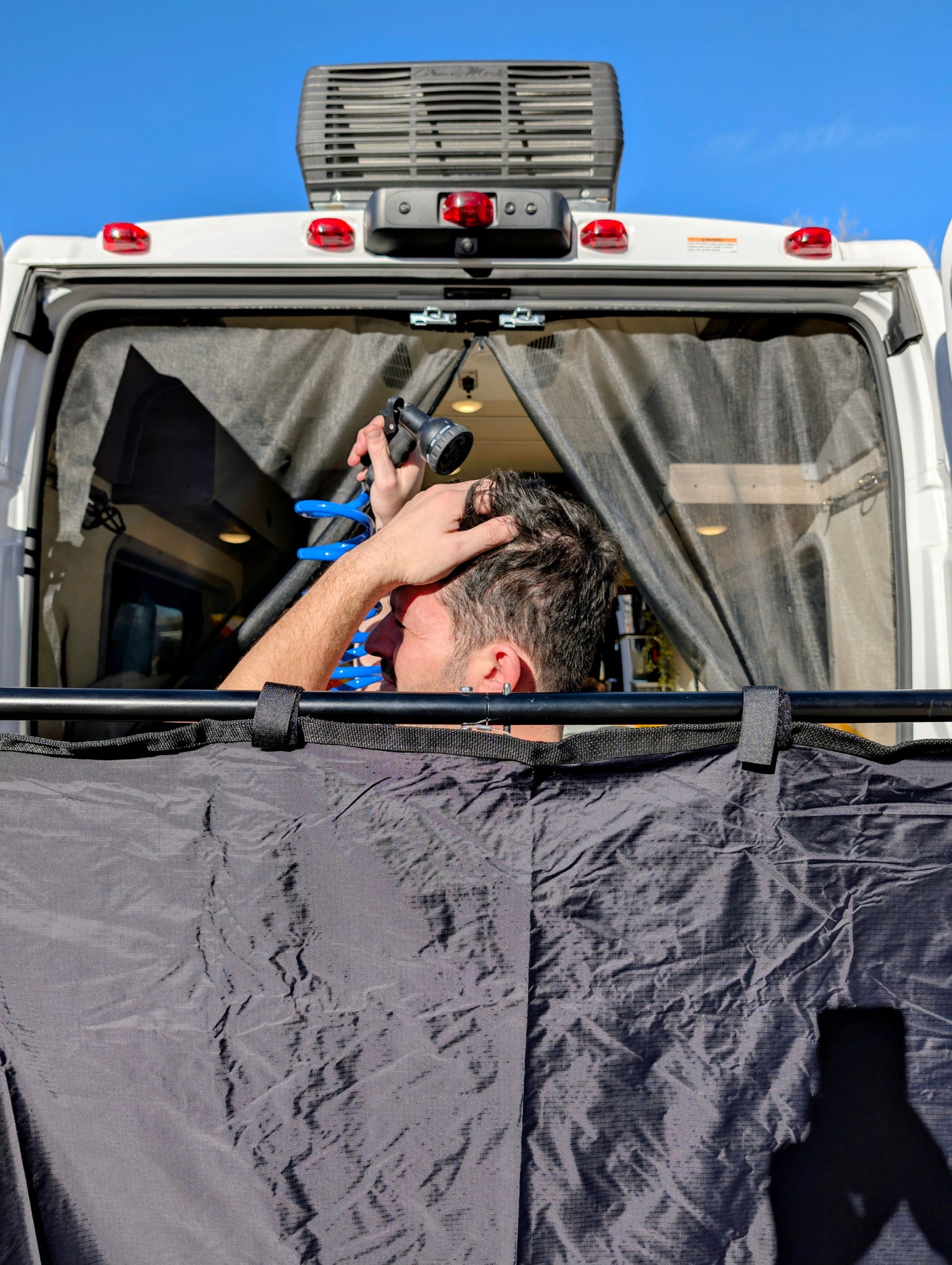 RV rental: Man showers with an outdoor hose from the back of a van, using a privacy screen for convenient camp hygiene.