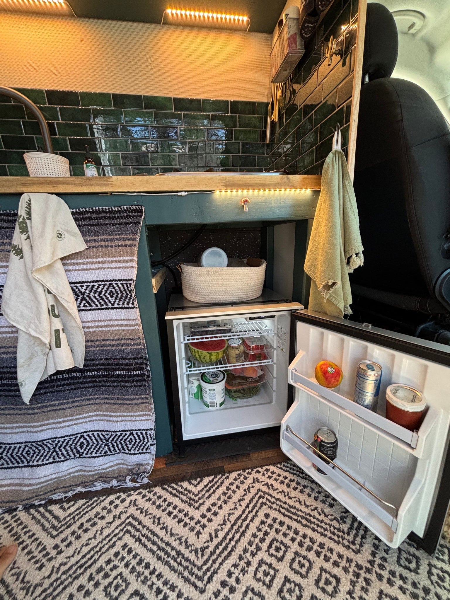 An RV rental interior features an open mini-fridge with food, green subway tile backsplash, strip lighting, and cozy patterned rugs.