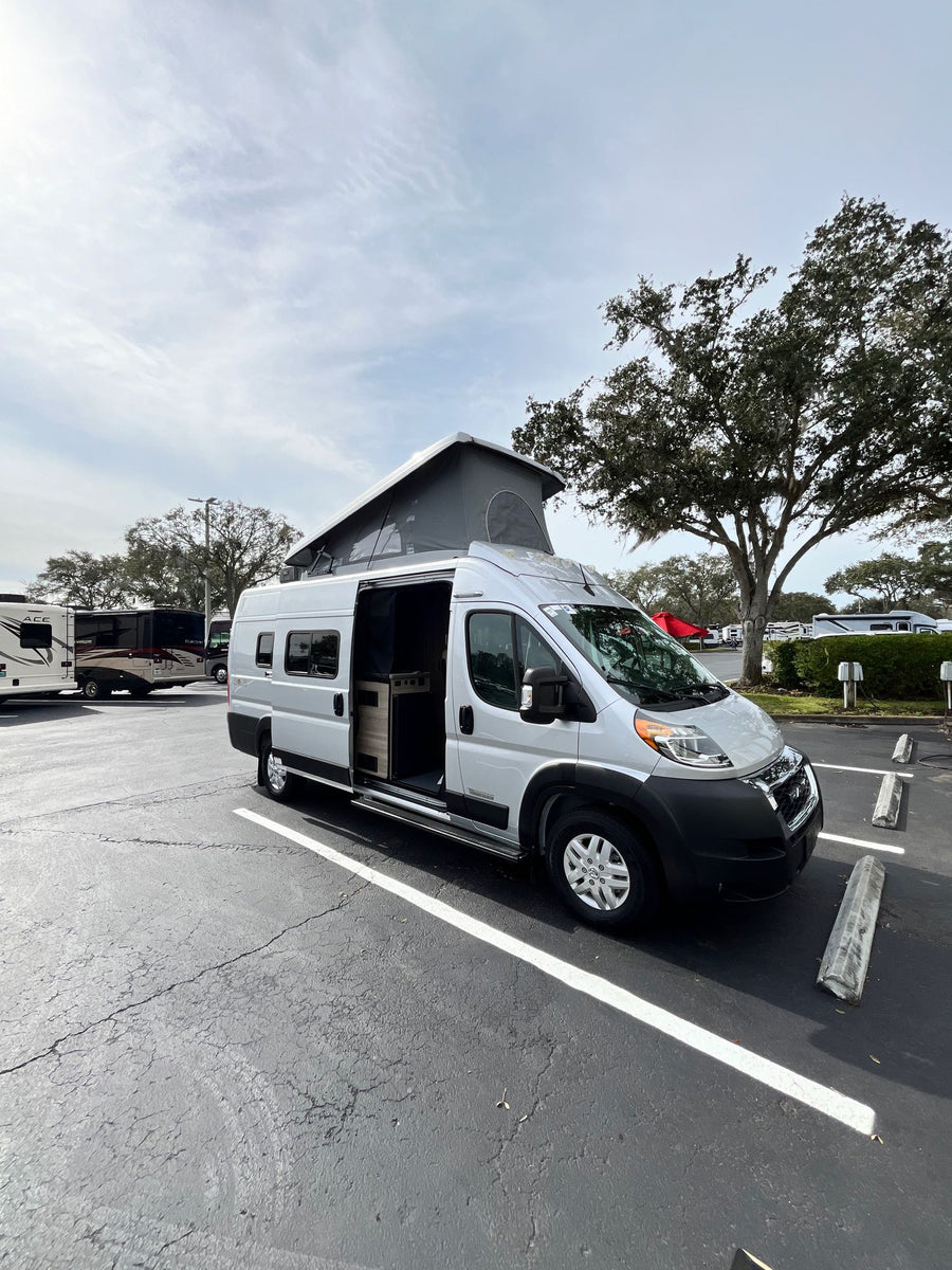 person's legs stretch on a comfy bed overlooking a sunset ocean.|Silver pop-top camper van with open side door