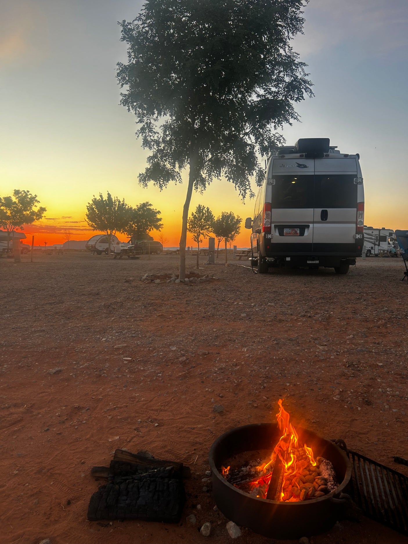 Vibrant sunset at an RV rental campsite. A silver RV is parked near a crackling campfire, offering a serene outdoor escape.