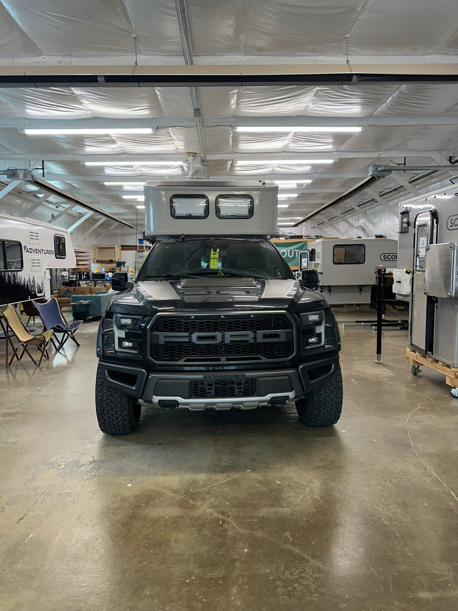 Black Ford Raptor truck with a light grey truck camper, available for rv rental, showcased in an RV manufacturing facility.