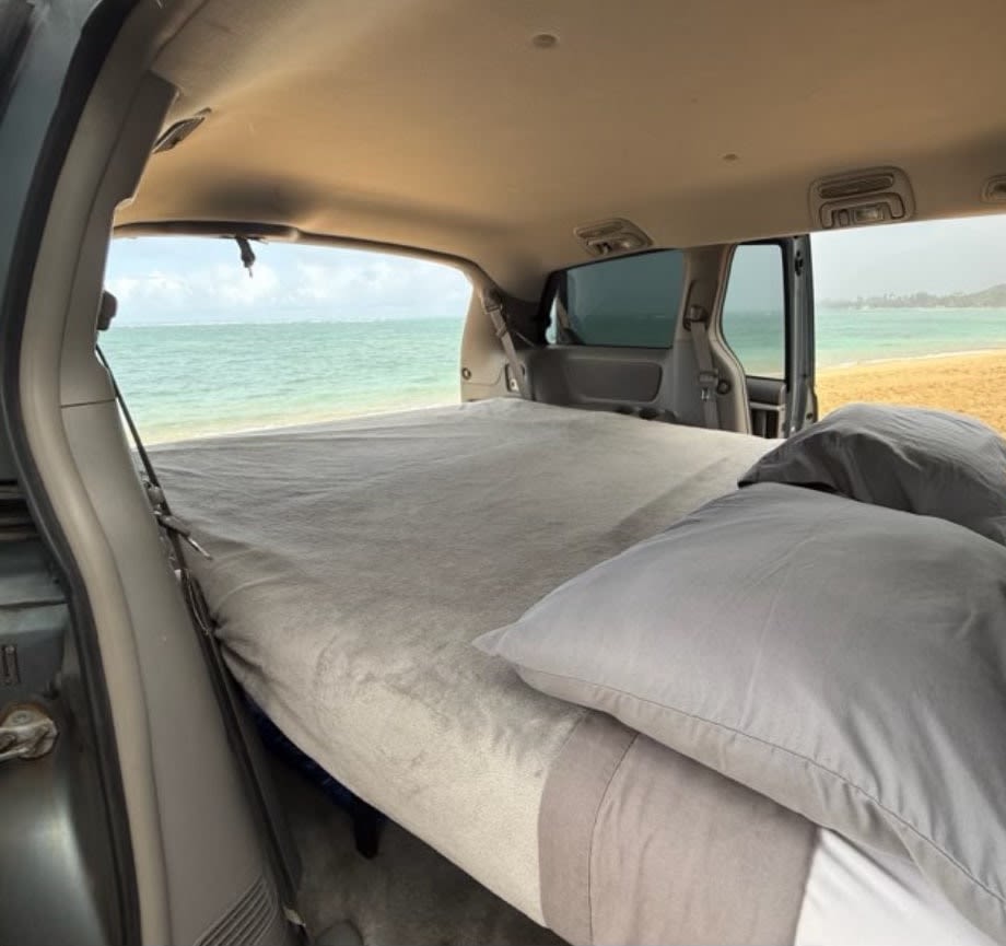 RV rental: Inside a customized van, a comfortable bed offers direct views of a sandy beach and blue ocean.