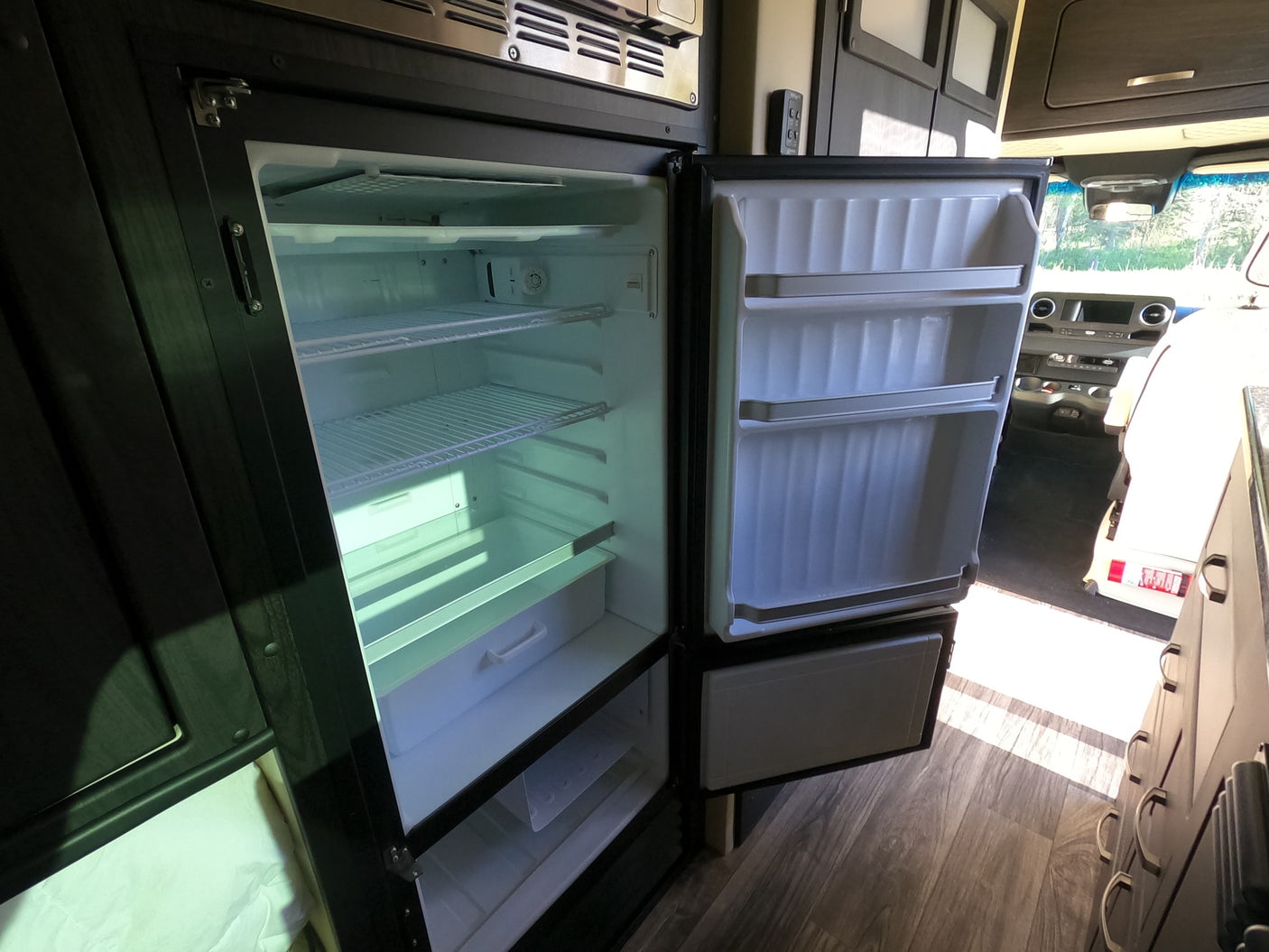 Empty, open two-door refrigerator/freezer inside an RV rental, showing internal shelves and door storage. Ready for your adventure.