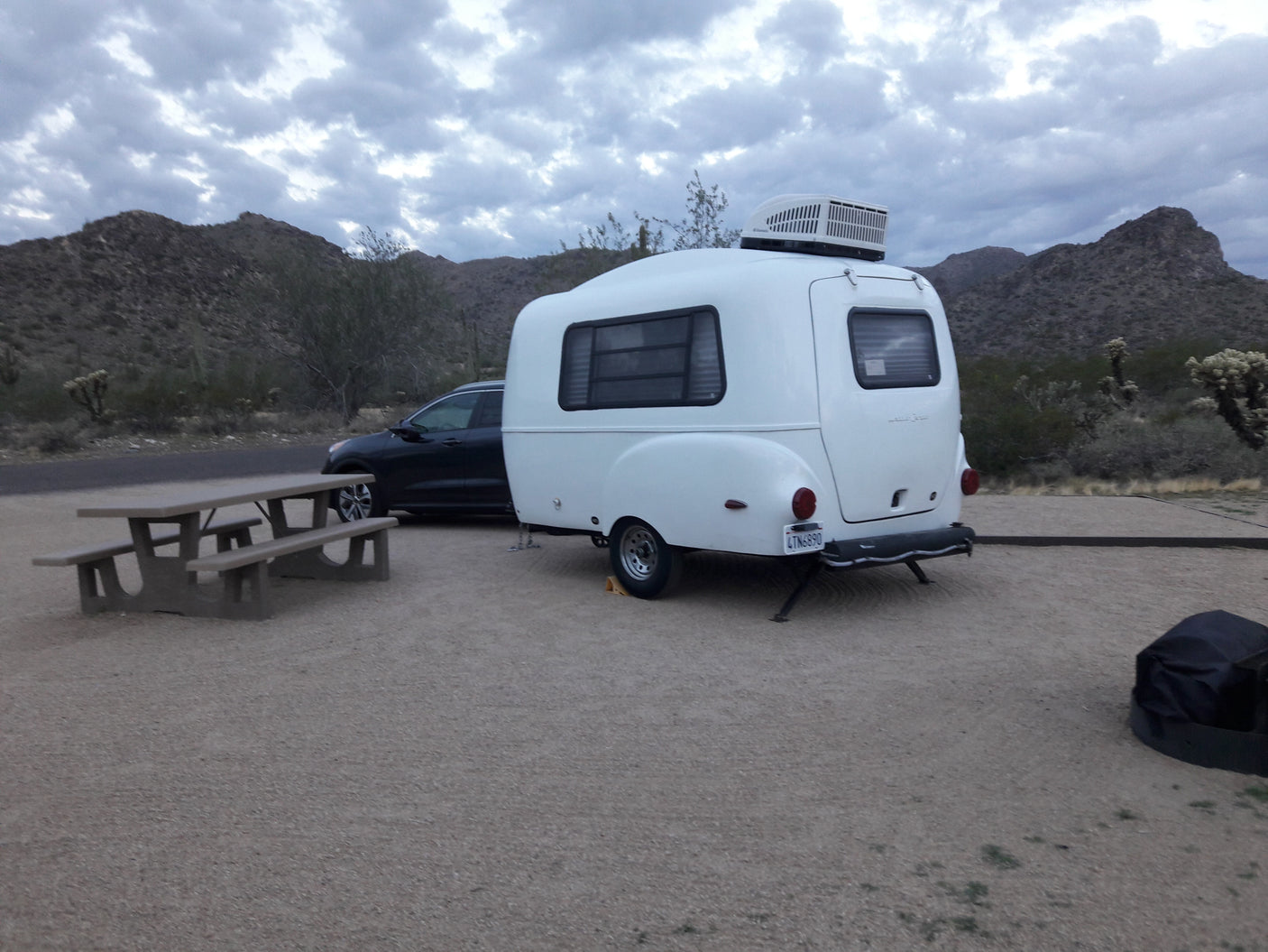 White RV rental & black SUV at desert campsite with a picnic table, mountains, and cloudy sky.